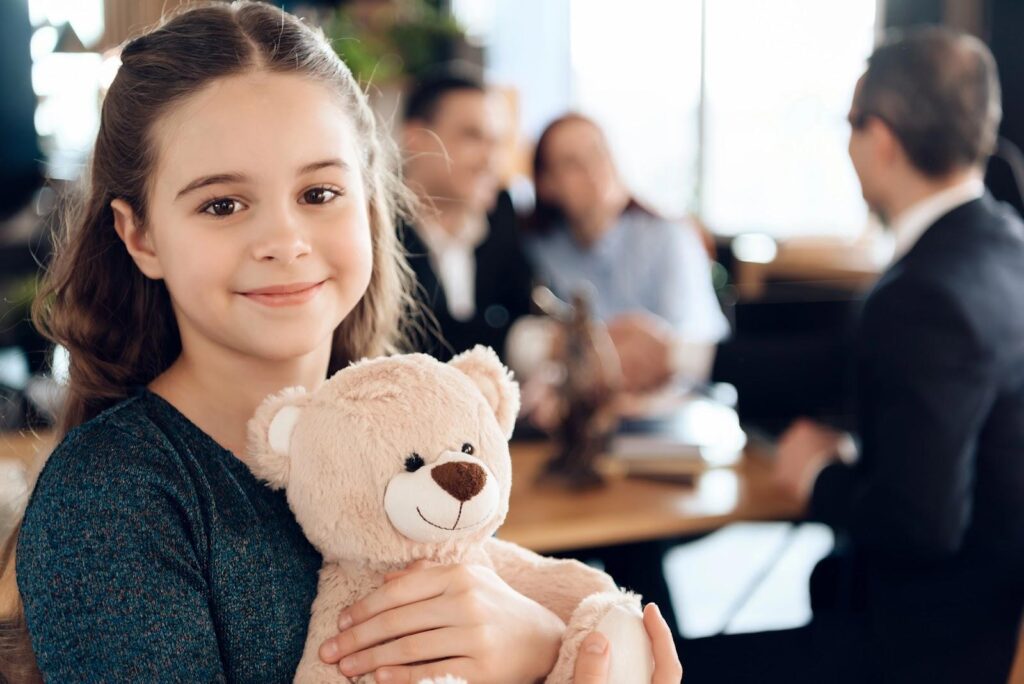 A little girl smiles and hugs a teddy bear as in the background, her parents work out custody issues during divorce mediation.