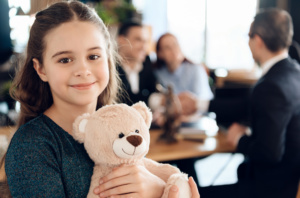 A child holding a teddy bear with parents talking to a family law attorney in the background.